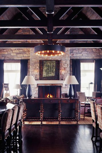 Living room with exposed beams, a round chandelier, a stone fireplace built into a rock wall, dark wood floor and a console table with two lamps with black bases