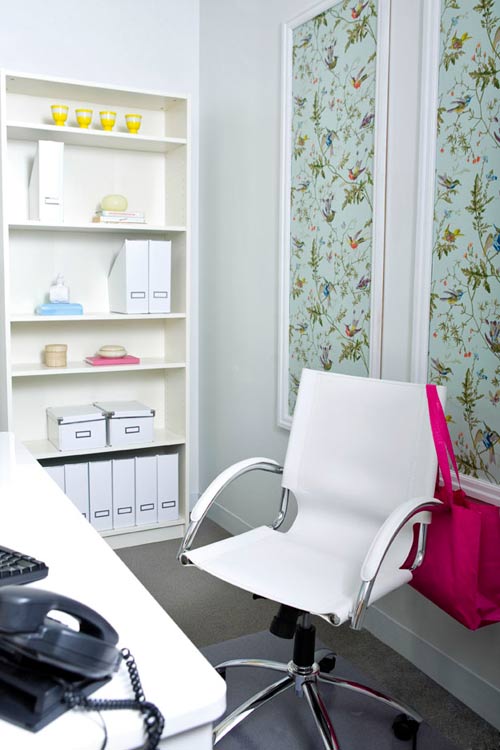 Office with framed light blue print with birds and foliage, a white rolling chair with a hot pink purse hanging off the back, and a white bookshelf with folders and shoeboxes