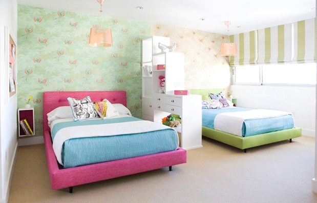 Girls bedroom with pink and green upholstered beds separated by a white drawer and shelf divider, each half of the room has the same wall paper in different color, the blinds are lime green striped 