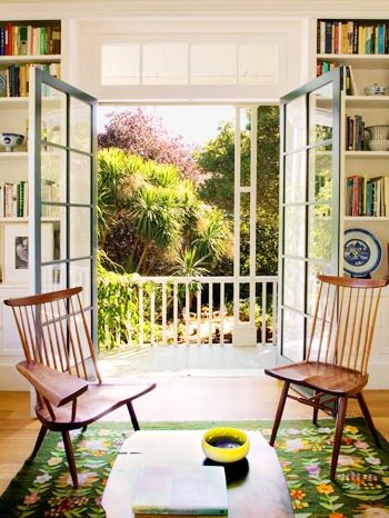 Living room with wood floor, built in bookshelves, french doors thrown open to a beautiful outdoor setting with two wood chairs facing each other on a green rug