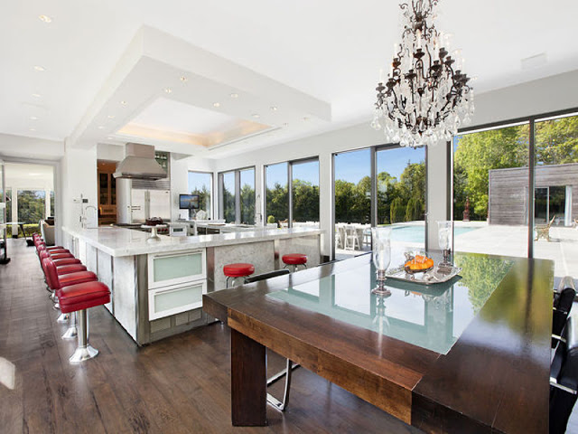 Open kitchen with red stools, wood floor, marble island counter tops, a wood table, chandelier and large windows overlooking a backyard with a pool