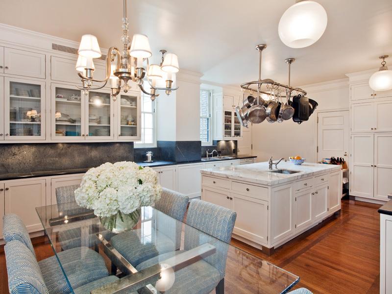 Eat in kitchen in a park avenue apartment with white cabinets, a pot rack hanging over an island with marble countertop, wood floors, and a glass table surrounded by blue upholstered seats with a chandelier above it