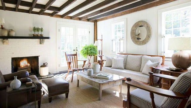 Living room with ticking stripe sofa chairs, exposed beams, a white fireplace, reclaimed wood coffee table, french doors with a sisal rug