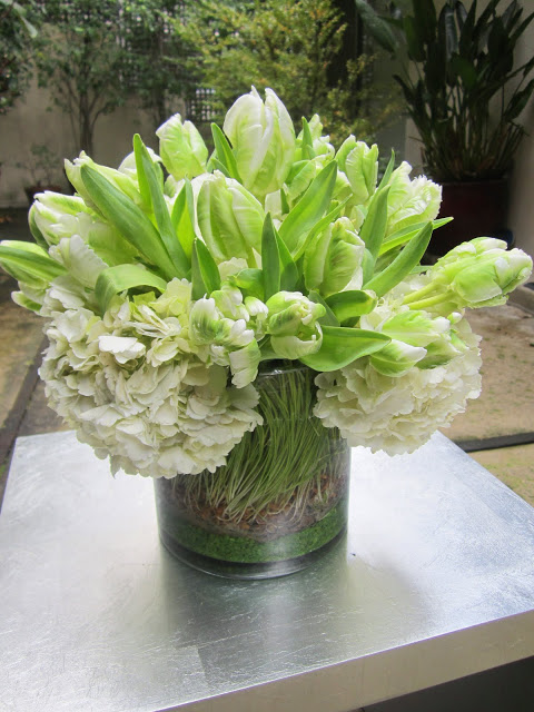 Green and white Parrot Tulips and Hydrangea in a glass cylinder vase with wheat grass and green sand on a metal table on a patio