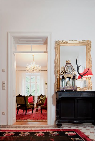Foyer in the Hammerhaus castle with a patterned tile floor, a black side table, a gold antique mirror and a view of a sitting room with wood floor and a large red carpet, three upholstered green chairs and a crystal chandelier