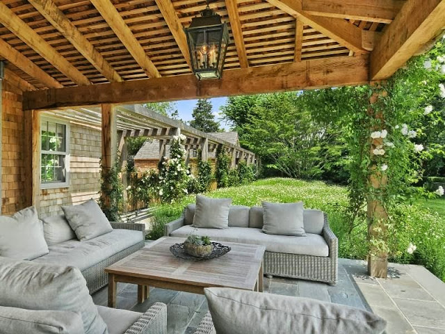 Patio of a house an East Hampton compound with grey sofas, wood table, a lantern light and a gorgeous yard