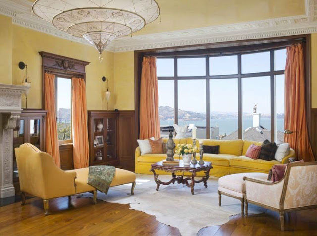 Sitting area in a san francisco mansion's master bedroom with yellow walls, a yellow sectional sofa, two armchairs with ottomans, wide wood paneled floor, animal hide rug, a wood coffee table, moulded ceiling, a fireplace and a large window with a view of the ocean. This window has floor length coral curtains