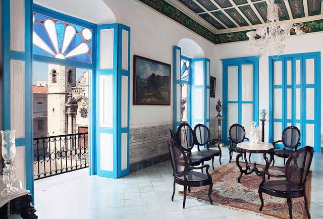 Sitting room in Cuban with blue trimmed panel doors, painted, paneled ceiling, a crystal chandelier and dark wood Louis XIV chairs arranaged around a small table, and a view from The Splendor of Cuba from Rizzoli