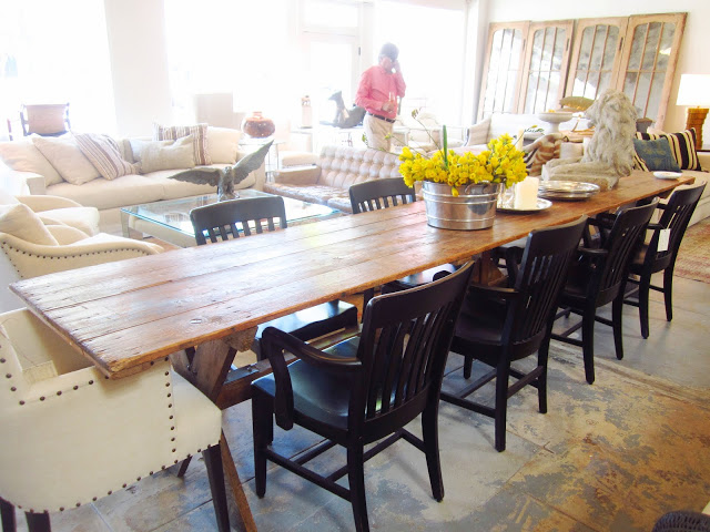 A long farmhouse trestle base dining table with a reclaimed wood top that seats at least 10 is surrounded by black wood chairs and an upholstered host chair with nailhead trim