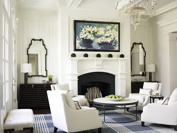Symmetrical living room with white armchairs, a round table with wire legs, a large blue and white plaid rug, a fireplace with a white molded mantel with a black chest of drawers and a glossy black mirror on each side