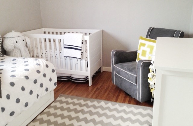 nursery with a white crib, glider upholstered gray armchair with white piping, and a yellow pillow