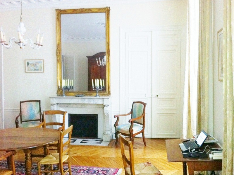 Dining room in a Paris apartment with a Carrara marble fireplace, a large mirror in a gold frame over the mantel, a white chandelier, a round wood table, herringbone wood floor, a small writing desk and a round pedestal wood table surrounded by wooden chairs