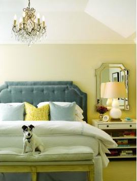 Bedroom with blue upholstered headboard, taupe walls, a crystal chandelier, a white nightstand, an arched vintage inspired mirror and an upholstered bench at the foot of the bed with a jack russell terrier sitting on it.