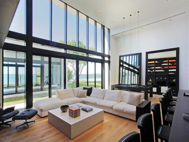 game room with wood floors, a white L shaped chair, two large black mirrors, and a square block coffee table. Instead of a traditional wall, they have a large glass wall with black panelling