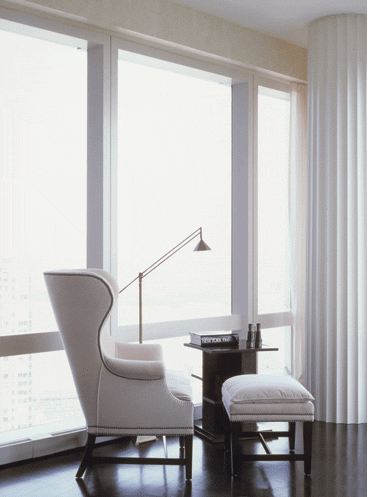 A white wingback chair and ottoman make for a sleek and stylish window seat in a city apartment with dark wood floor and wall to ceiling windows