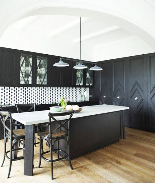 Kitchen with dark cabinets with diamond shaped molding details, light rustic looking floors, a very bold trellis pattern black and white backsplash and glossy black counters. The island in the center has a glossy white counter top.