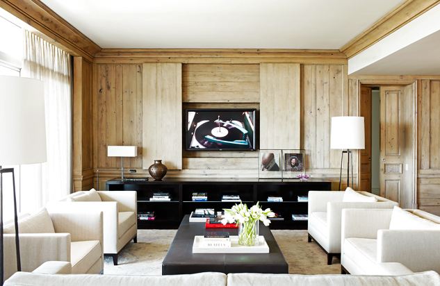 Modern family room with dueling white armchairs, a long dark ottoman being used as a coffee table, black book shelves and light wood paneling concealing a television