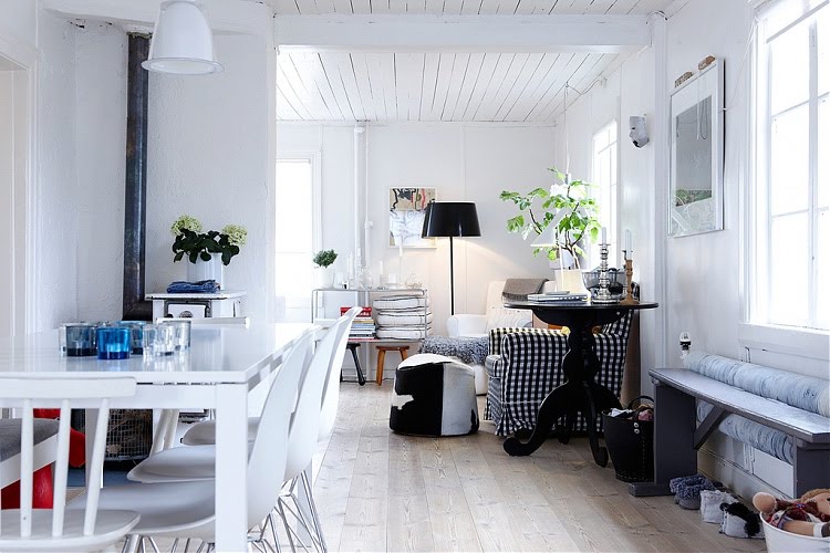 White living room with gingham covered armchair, black floor lamp, black and white piped floor cushions, round black table, a light blue bench under a window and a white table with white eames chairs