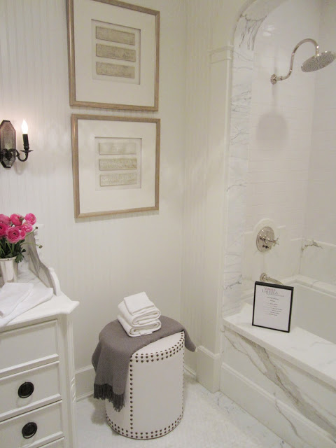 Bathroom in the House of Windsor with white beadboard paneled walls, carrara marble bath with an arch, a little leather stool with nail head trim, white cabinets, pink peonies and a wall mounted candlestick inspired light 
