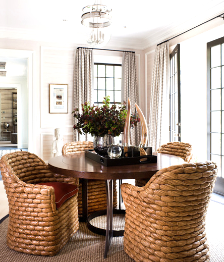 breakfast nook with four chairs made of a large wicker weave surrounding a wooden table with a serving tray holding a vase of flowers, a glass ball and a pair of horns. The room is lit by large black framed windows with floor length patterned curtains