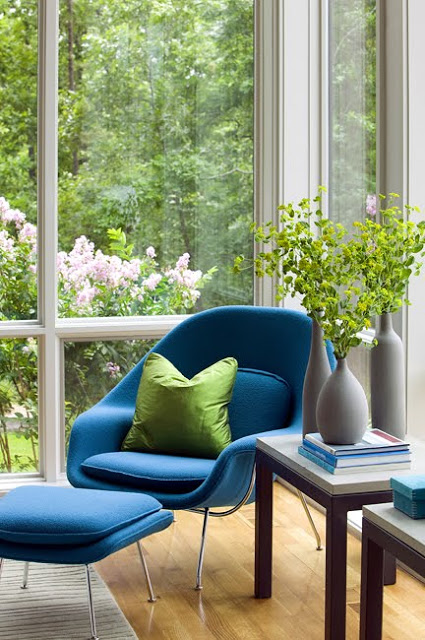 Room with floor to ceiling windows with a garden view, a blue Saarinen womb chair, hard wood floors and two side tables holding bottles of flowers and books