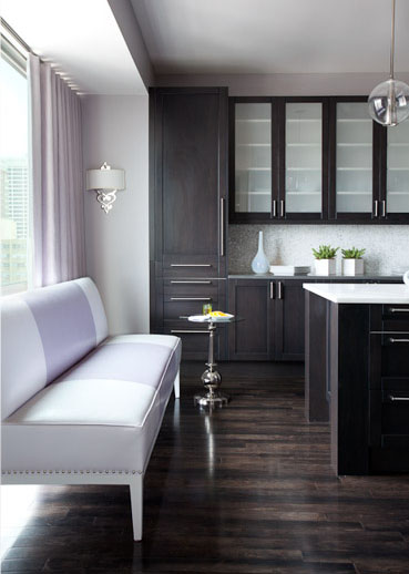 Alternative view of the kitchen with dark wood floors and cabinets from floor to ceiling. In this view you can see the picture window with lavender floor length curtains and a long lavender upholstered bench with nail head trim