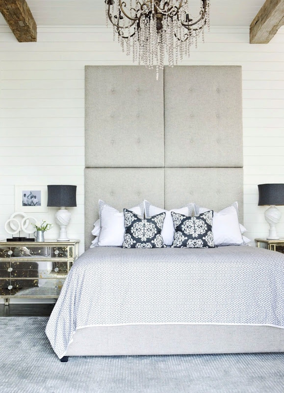 Bedroom with white beadboard walls and exposed beams, high ceiling, a tall, tufted grey headboard, mirrored side table, a grey rug and a crystal chandelier