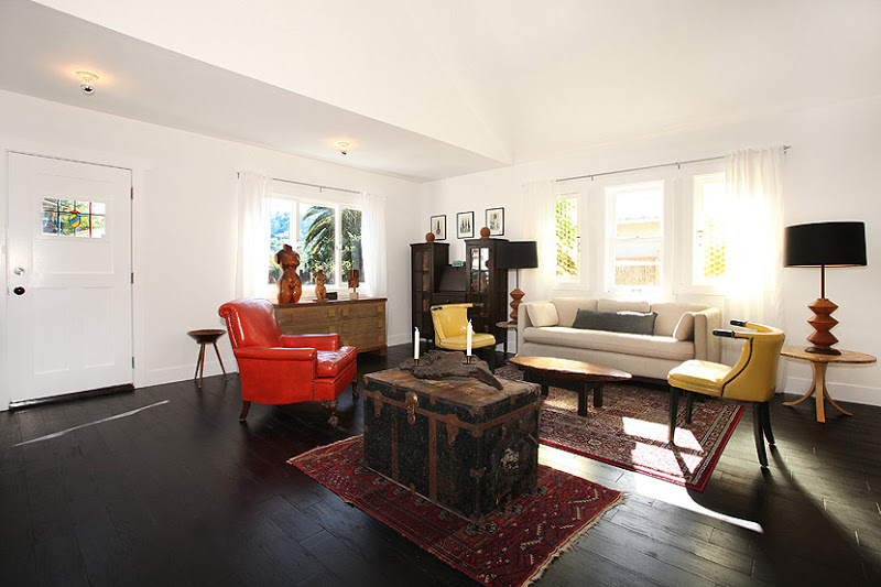 Alternative view of the living room in a cottage with dark wood floor, yellow settees, a taupe armchair, a black writing desk, a vintage chest holding a piece of polished wood, two Turkish rugs and a wooden chest of drawers with sculptures of the female figure, and a red leather armchair