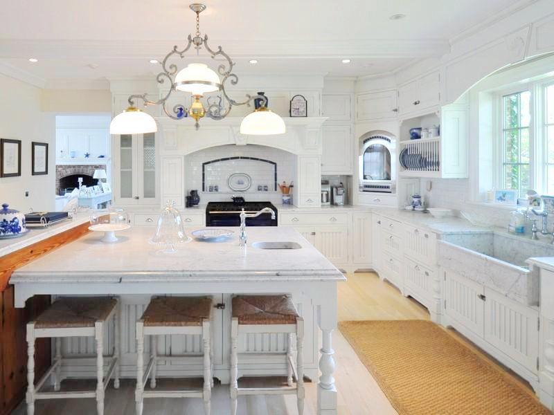 Kitchen with white cabinets, light wood floor, a white island surround by white stools with sea grass chairs, a farmhouse sink, a chandelier and a sea grass rug
