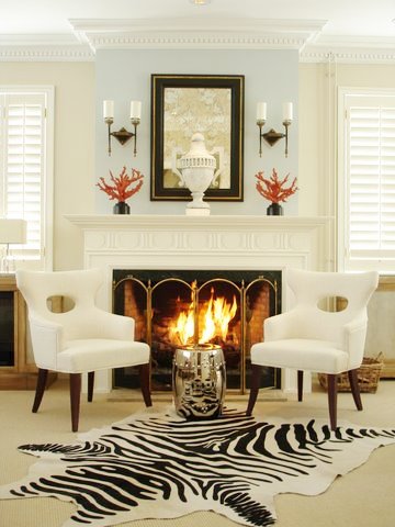 Living room in Ron van Empel of Empel Collections' home with zebra rug, a silver garden stool, fireplace with paneled wood mantel and a pale blue wall, and two white side chairs