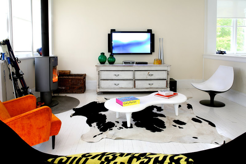 Ecletic family room by Robert and Cortney Novogratz with cow hide rug, black bow tie sofa, white modern coffee table, orange armchair and freestanding fireplace