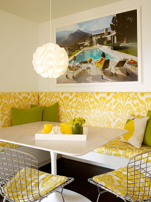 Yellow and white breakfast nook with built in banquette seating with ikat fabric on the seats, a Le Klint pendant light, Bertoia chairs and a white rectangular table with a Saarinen base