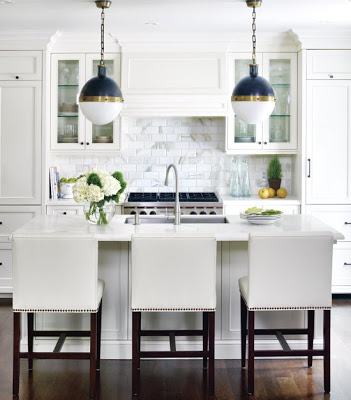 White kitchen with marble subway tile backsplash, cabinets topped with crown molding, a custom panel to cover range hood, nailhead trim counter stools and Thomas O'Brien antique brass, blue and white glass banded Hicks pendant lights