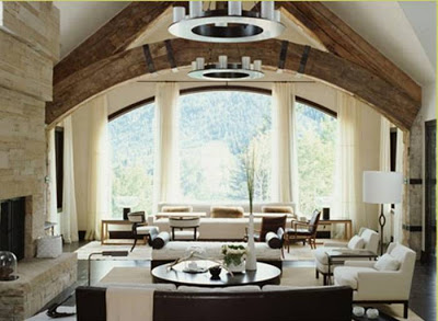 Living room in a lodge with a huge fireplace, beamed ceilings, two sofa, a large lounge chair, a few modern lamps, chandeliers and dark wood floor
