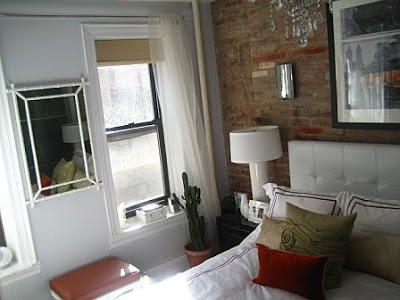 Bedroom after remodeling with white upholstered headboard, bedding with red embroidery, exposed brick wall and a stool