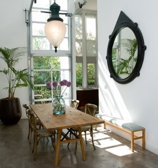 Dining room in a loft designed by Nickey-Kehoe with a round black mirror on the wall, traditional farmhouse style table and wood chairs and a large potted palm 