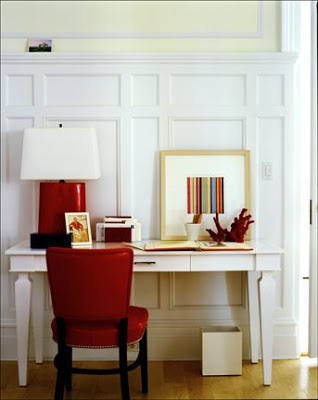 Home office with white paneled walls, white desk, a red leather chair, table lamp and red and black accessories