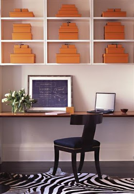 Home office by Sara Story with a large wood slab wall mounted desk, zebra rug, empire style chair and Hermes orange storage boxes in white open shelves
