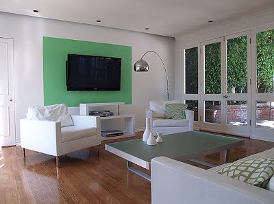 Modern dining room designed by Vanessa de Vargas with white leather armchairs and sofa, frosted glass and brushed metal coffee table, French doors with a view of a lush patio and a green color block on one wall with a flat panel TV