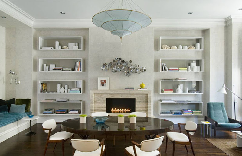Grey dining room with white shadow box shelving, dark brown round wood table surrounded by retro white leather and wood chairs, a blue umbrella style chandelier and velvet cushion on a window seat and a silver 1950s inspired wall sculpture over the fireplace mantel