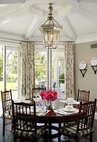 Elegant dining room with tan walls, tolie print curtains, floor to ceiling French doors and paned windows, Queen Anne dining chairs with striped set cushions and a brass lantern style pendant light