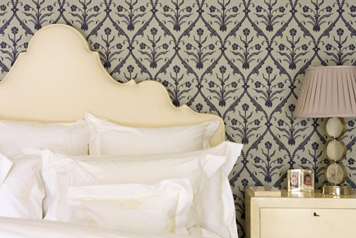 Bedroom with a white Queen Ann headboard, floral print blue and grey wallpaper. and a brass lamp with a pink shade on a cream colored nightstand