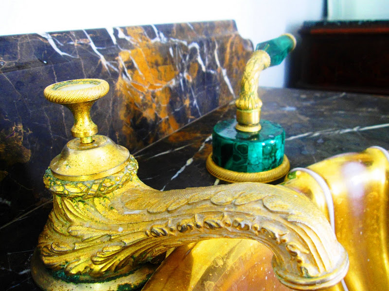Close up of the sink fixtures which include a gold faucet featuring a leaf design on it and handles that adorned with emerald green malachite trim in a master bathroom