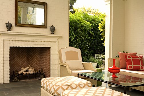 Loggia with a white washed brick fireplace, red accent pillows on off white wicker furniture and a red ceramic vase on a glass coffee table