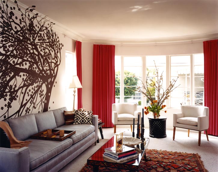 Living room with a grey sofa, African Bamileke side table, a huge wall decal of a tree and bright red curtains