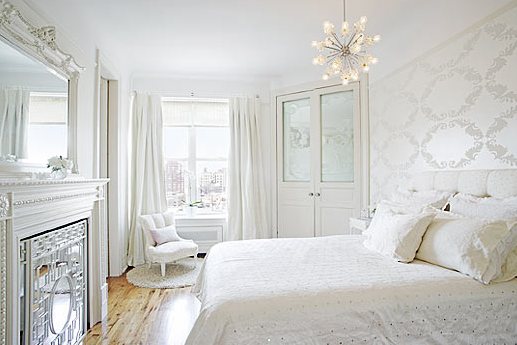 White bedroom by Kelly Giesen with a tufted headboard, patterned wallpaper, chandelier and a mirrored fireplace