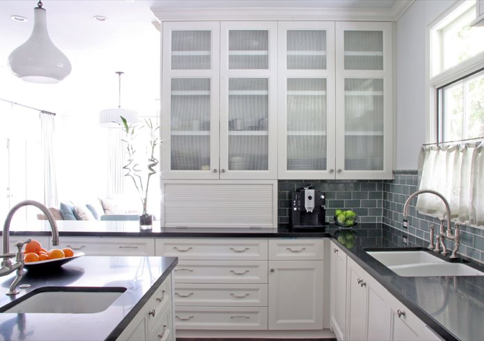 White kitchen with cabinets with recessed panel doors and drawers, reeded glass upper cabinets, slate subway tile backsplash, undermount sinks and dark stone countertops