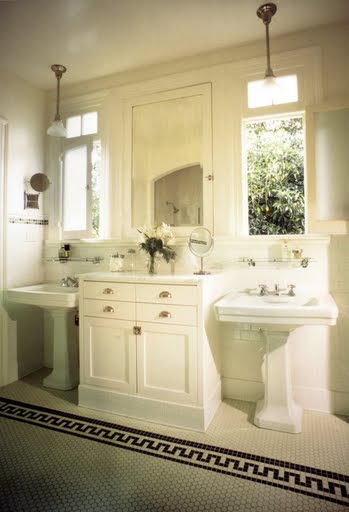 Classic white bathroom by Kevin Oreck with two pedestal sinks under windows flanked by built in bathroom cabinets and a white tile floor with a black border