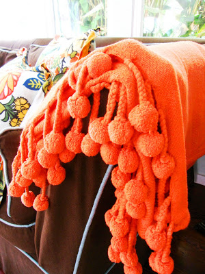 Bright orange throw with pompoms from Calyspo Home on a brown sofa with turquoise piping