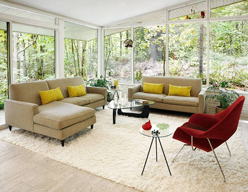 Modern living room with floor to ceiling windows, two taupe sofas with yellow accent pillows, a Noguchi glass coffee table and a red Eero Saarinen Womb Chair